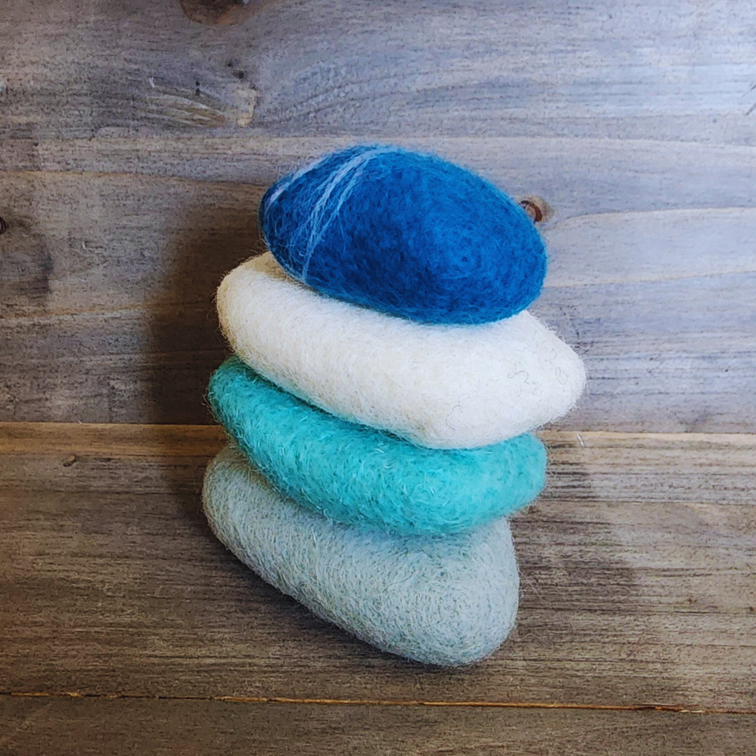 A slightly angled view of four handmade wet-felted Zen Stones stacked, showing textures and colors of light gray, teal, white, and a deep blue with white striations, on a rustic wooden background.