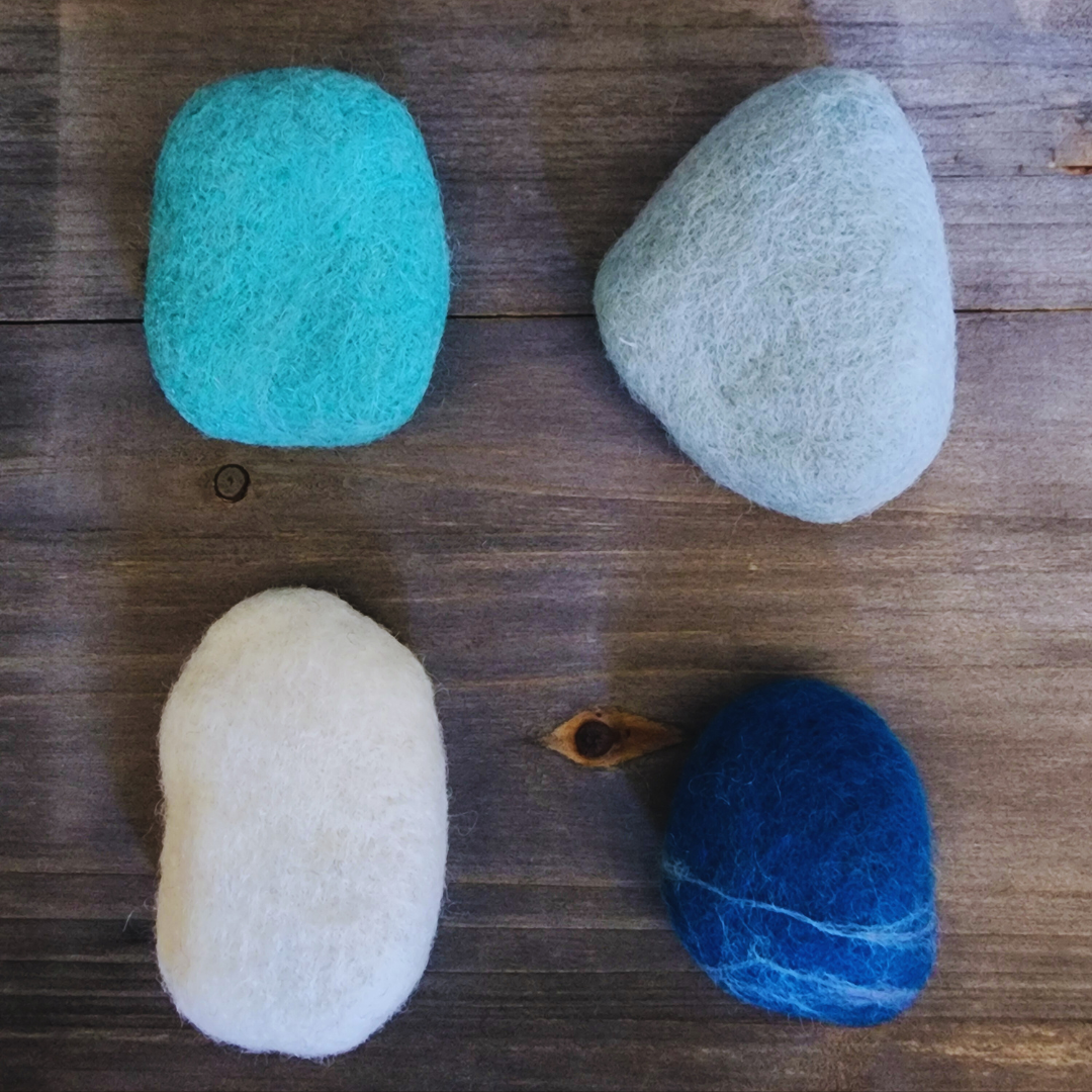 Four handmade wet-felted Zen Stones in an assortment of blue and white tones (teal, light gray, white, and deep blue with white striations), arranged unstacked on a wooden surface.
