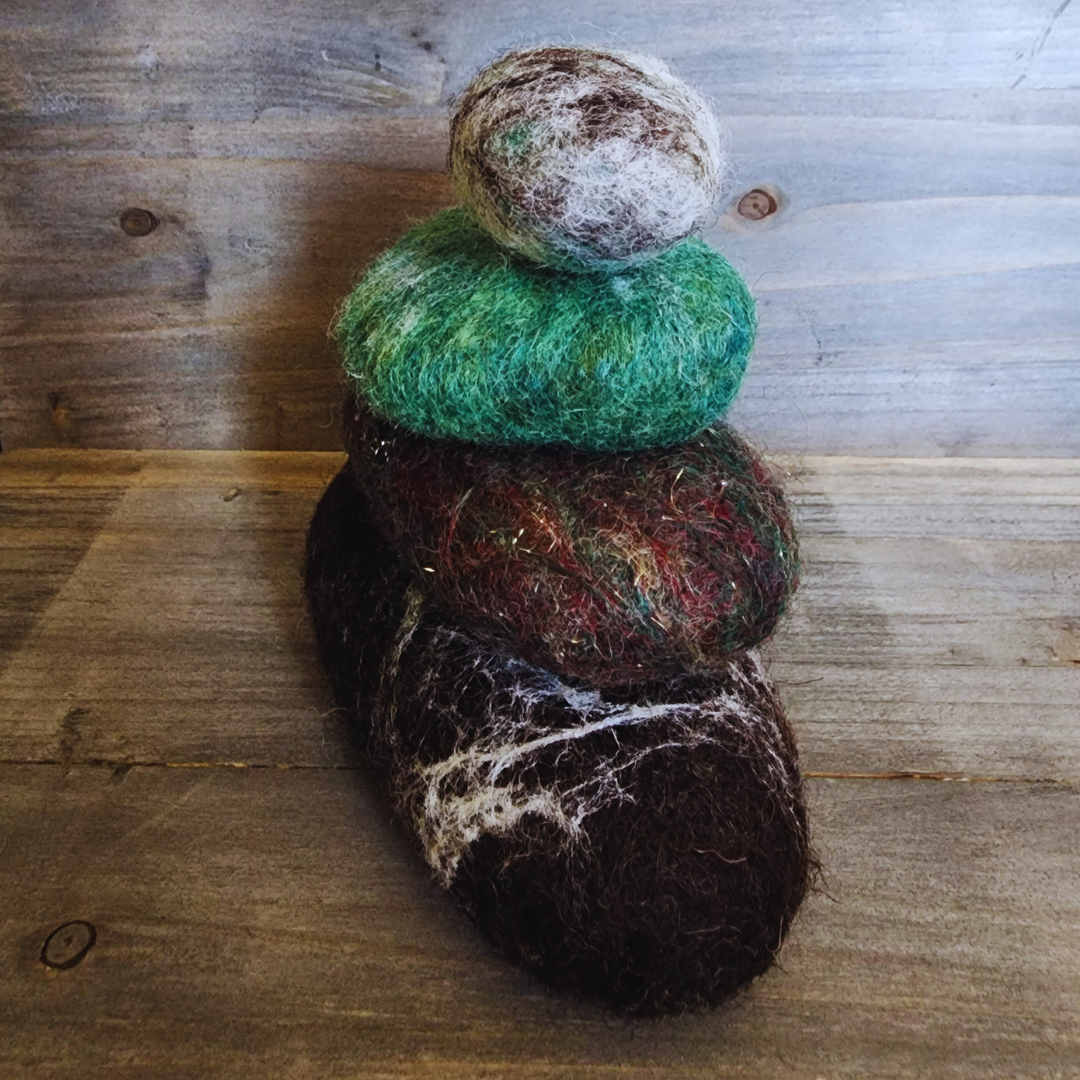 Four handmade wet-felted Zen Stones stacked vertically, from bottom to top: dark brown with white marbling, mottled green/red with gold flecks, vibrant green, and a mixed brown/gray/white top stone, on a wooden surface.
