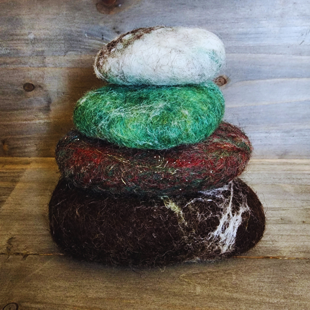 Four handmade wet-felted Zen Stones stacked in a neat vertical column, showing colors of dark brown with white marbling, mottled green/red with gold flecks, vibrant green, and a light gray/brown top stone, against a rustic wooden background.