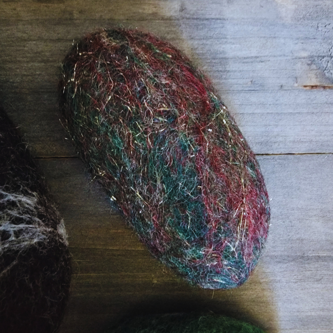 A close-up overhead view of a single handmade wet-felted Zen Stone, mottled with dark red, green, and hints of gold/silver sparkle, resting on a wooden background.