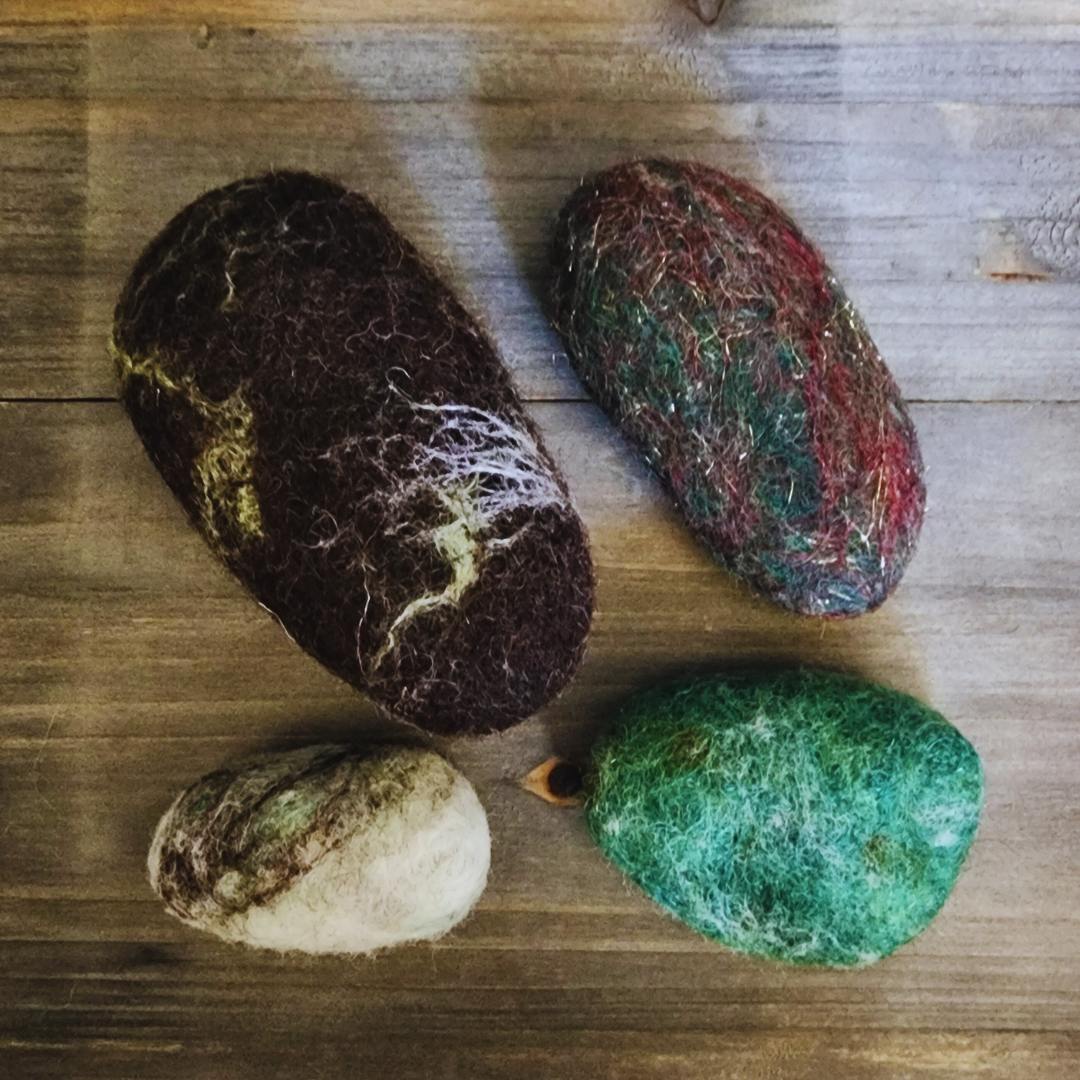 Four handmade wet-felted Zen Stones in earthy tones arranged unstacked on a wooden surface: a large dark brown with white marbling, a mottled green/red with gold flecks, a small light gray with dark swirls, and a vibrant green.