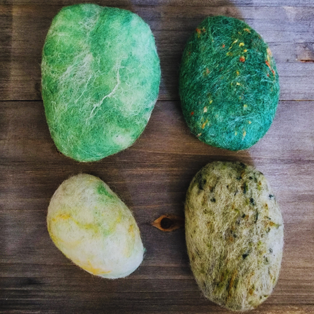 Four handmade wet-felted Zen Stones in various shades of green, yellow, and white, arranged unstacked on a wooden surface.
