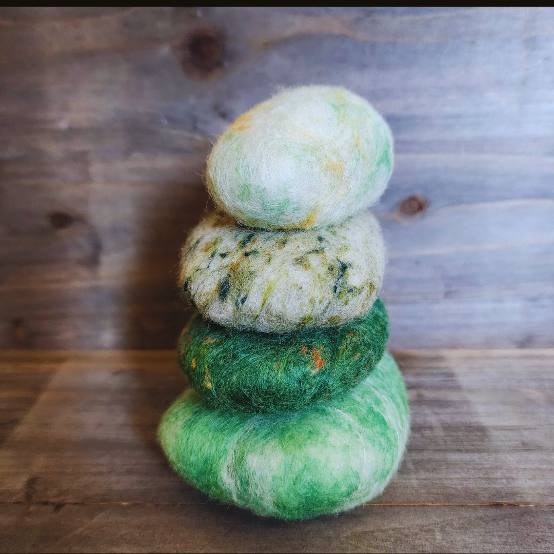 Four handmade wet-felted Zen Stones stacked vertically, from bottom to top: light green with white marbling, dark green with orange/red flecks, mottled light green with dark specks, and a light green/white with yellow accents, on a wooden surface.