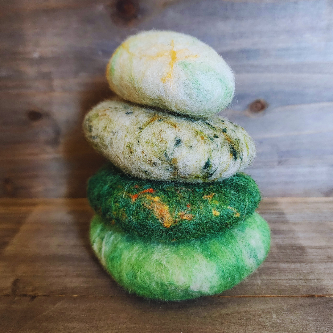 A close-up view of four handmade wet-felted Zen Stones stacked in a neat vertical column, showing shades of light green with white marbling, dark green with flecks, mottled light green, and a light green/white with yellow, on a rustic wooden background.