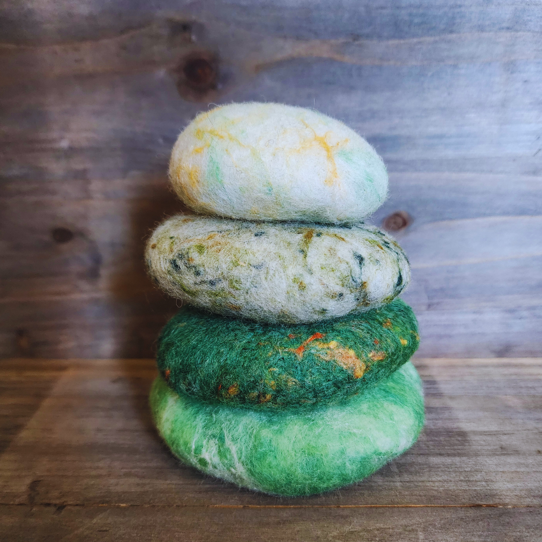 Four handmade wet-felted Zen Stones stacked vertically, featuring shades from bottom to top: light green with white marbling, dark green with orange/red flecks, mottled light green with dark specks, and a light green/white with yellow accents, on a wooden background.