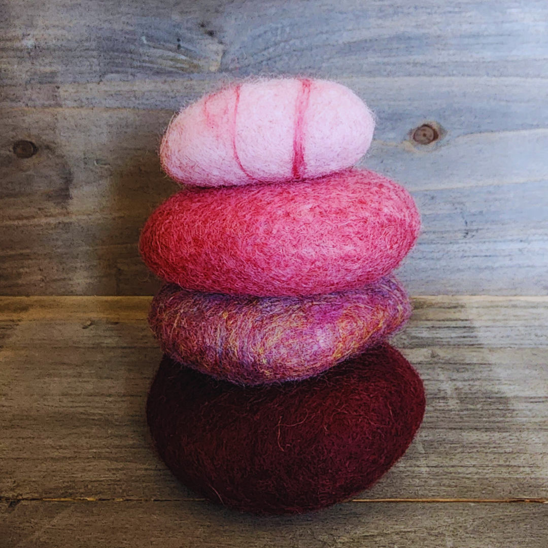 Four handmade wet-felted Zen Stones stacked vertically, featuring colors from bottom to top: dark red-purple, mottled pink and purple, bright pink, and light pink with red stripes, on a wooden background.