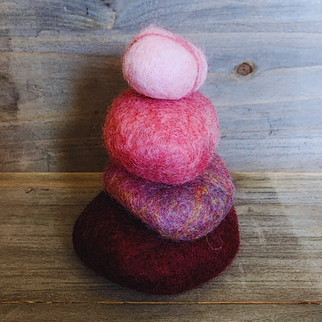 Four handmade wet-felted Zen Stones stacked, with the top stone being a solid light pink and the bottom three mirroring the dark red-purple, mottled, and bright pink tones, on a rustic wooden background.