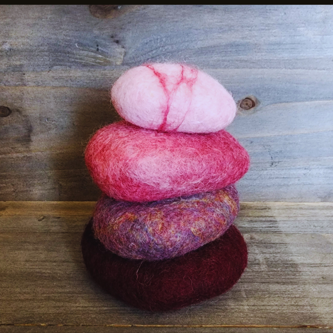 Four handmade wet-felted Zen Stones stacked vertically, showing a bottom dark red-purple, a mottled pink/purple, a bright pink, and a top light pink with red stripes, on a wooden background.
