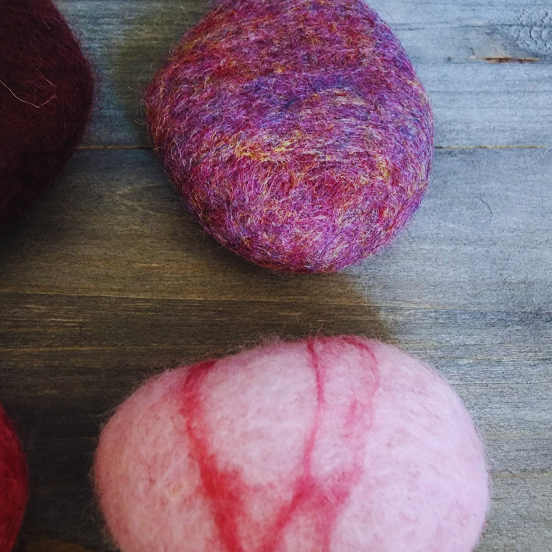 A close-up overhead view focusing on two handmade wet-felted Zen Stones, one a mottled pink and purple color, and the other a light pink with distinct red stripes, on a wooden surface.