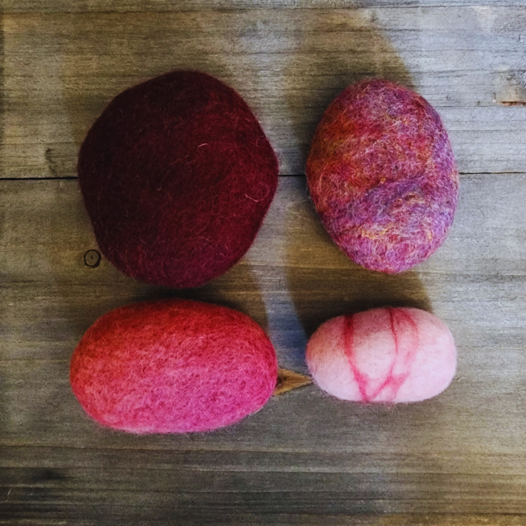 Four handmade wet-felted Zen Stones in shades of pink and purple, arranged unstacked on a wooden surface: a dark red-purple, a mottled pink and purple, a bright pink, and a light pink with red stripes.