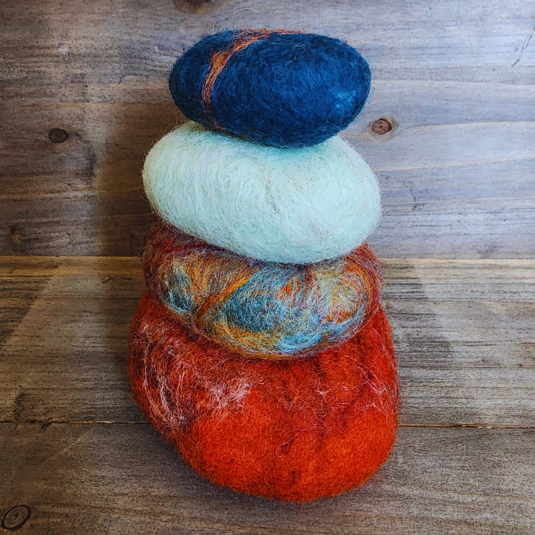 Four handmade wet-felted Zen Stones stacked vertically, displaying earthy and sky colors: a reddish-orange, a multi-colored stone with orange, blue, and brown, a soft light blue, and a dark blue with orange streaking, on a wooden surface.