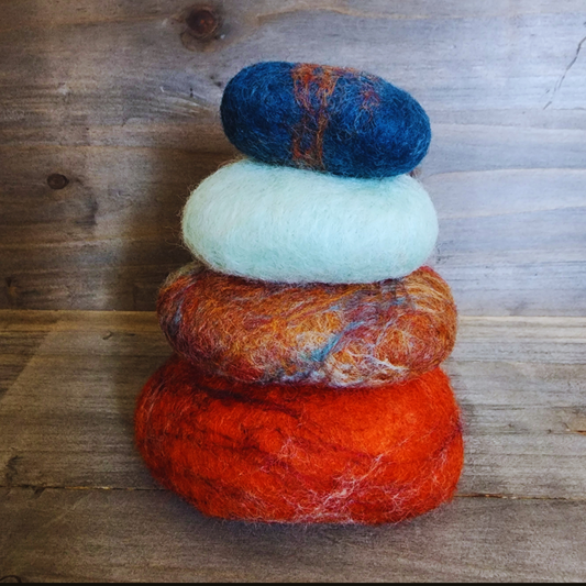 Four handmade wet-felted Zen Stones stacked in a neat vertical column, from bottom to top: deep orange, mottled orange/brown/blue, light turquoise, and a deep blue with orange streaks, on a wooden background.