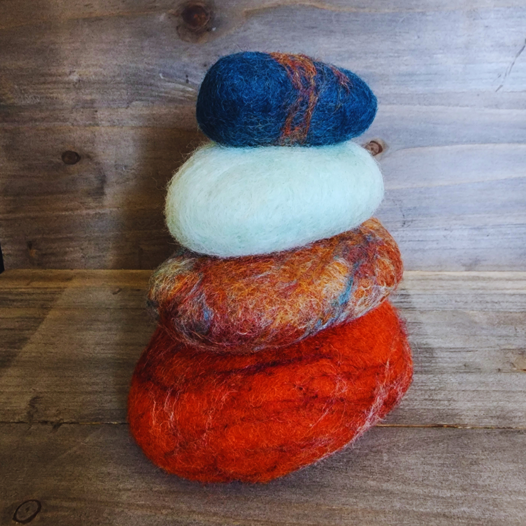 Four handmade wet-felted Zen Stones stacked vertically in colors reminiscent of Sedona: deep orange, mottled orange/brown/blue, light turquoise, and a deep blue with orange streaks, on a wooden surface.