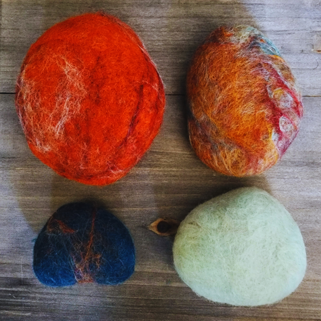 our handmade wet-felted Zen Stones arranged unstacked on a wooden surface, showing vibrant colors: a large solid orange, a mottled orange/brown/blue, a small dark blue with red streaks, and a light turquoise.