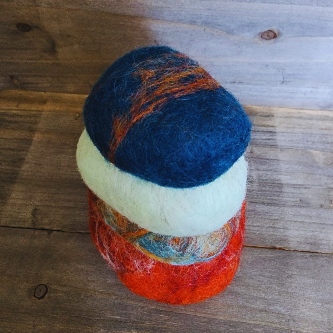 A slightly angled view of four handmade wet-felted Zen Stones stacked, showing shades of red-orange, mottled earthy tones, light teal, and a dark blue with hints of orange, on a rustic wooden background.