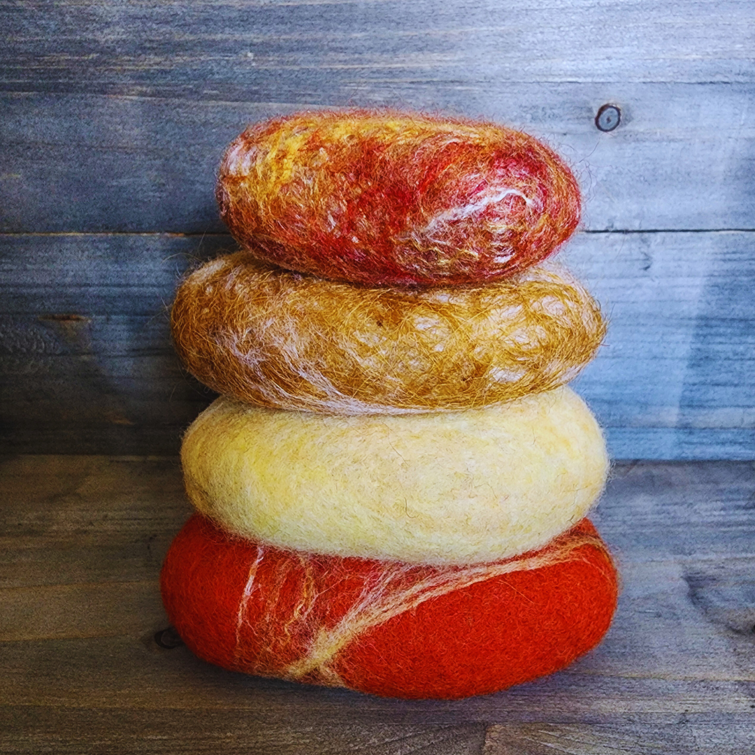 Four handmade wet-felted Zen Stones stacked vertically, from bottom to top: red-orange with white and yellow streaks, light yellow, golden brown with white, and mottled orange and red, on a wooden background.