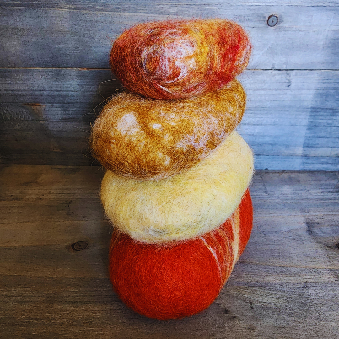 Four handmade wet-felted Zen Stones stacked vertically in warm tones, displaying colors of red-orange, light yellow, golden brown with white, and a mottled top stone with orange and red, on a wooden surface.
