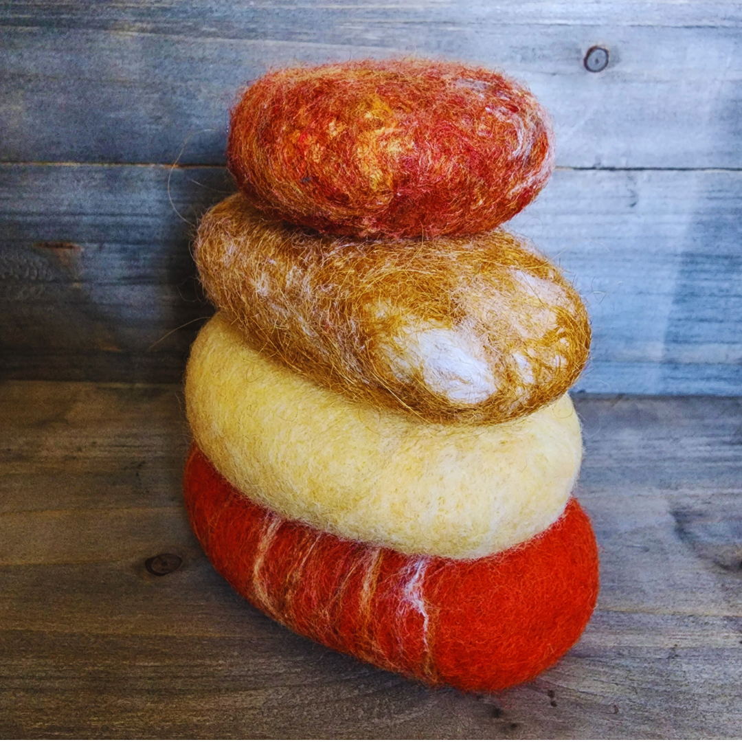 Four handmade wet-felted Zen Stones stacked vertically, featuring warm colors: red-orange, golden yellow, mottled golden brown, and a top stone with mixed orange and red, on a rustic wooden background.