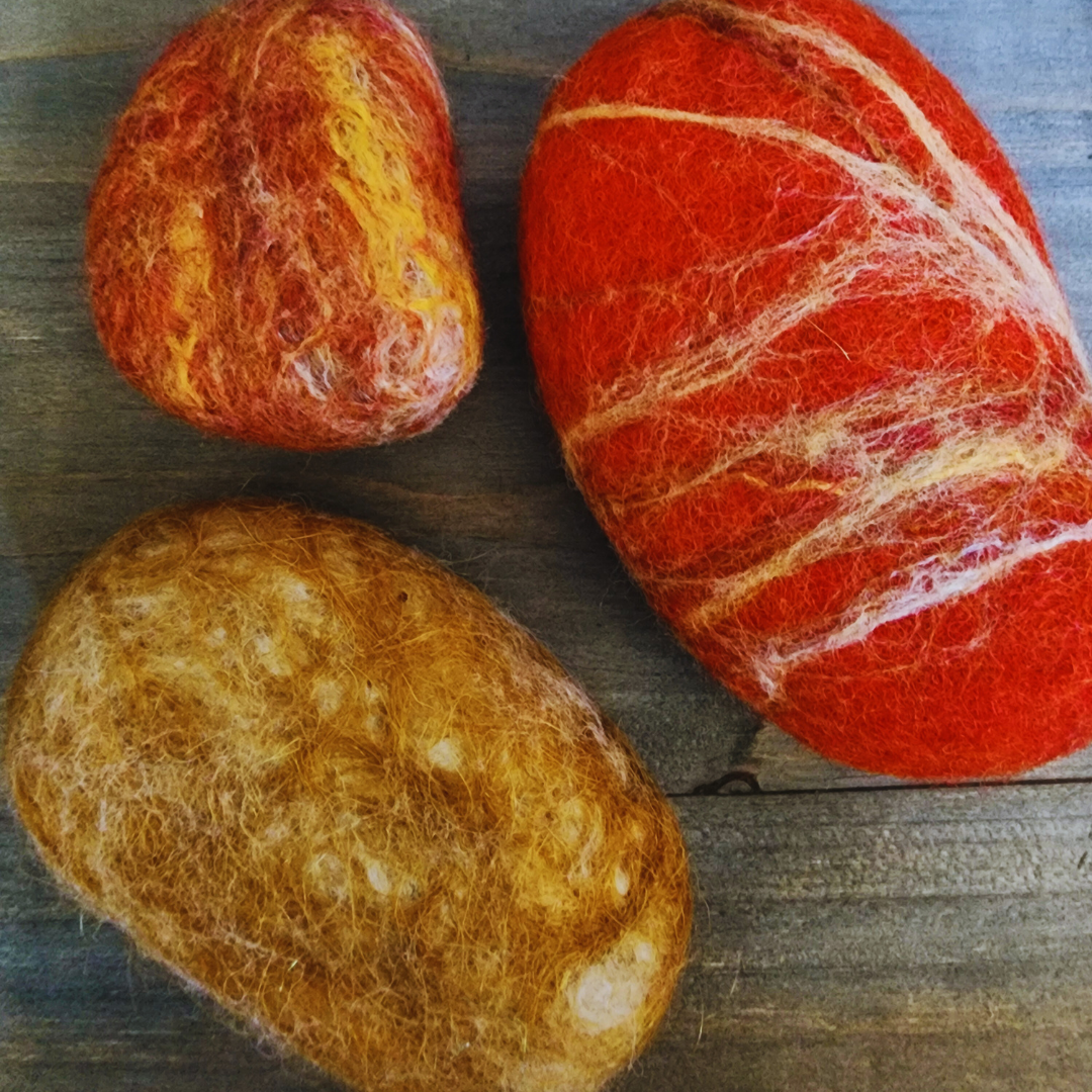 Three handmade wet-felted Zen Stones in warm, sunset-inspired colors (mottled orange and yellow, solid orange with white and yellow streaks, and golden brown), arranged unstacked on a wooden surface.