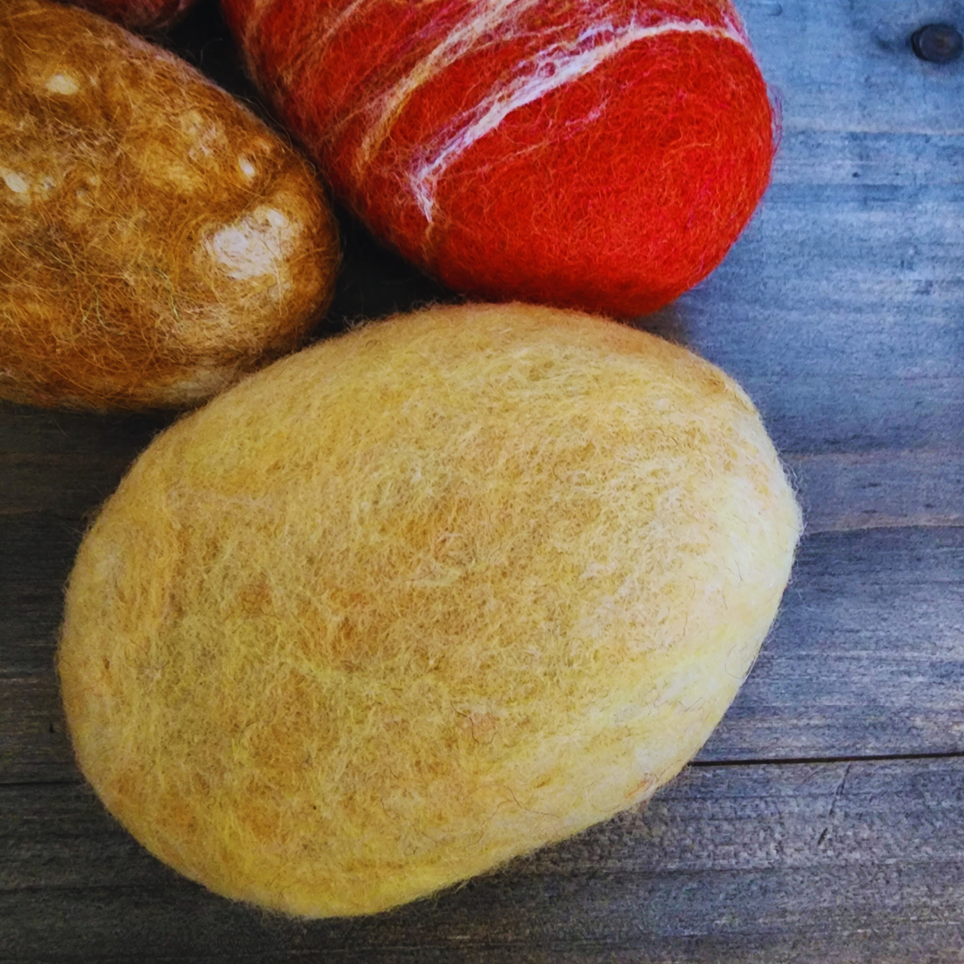 A close-up overhead view of two handmade wet-felted Zen Stones, one solid light yellow and the other a vibrant red-orange with white and yellow streaks, resting on a wooden background.
