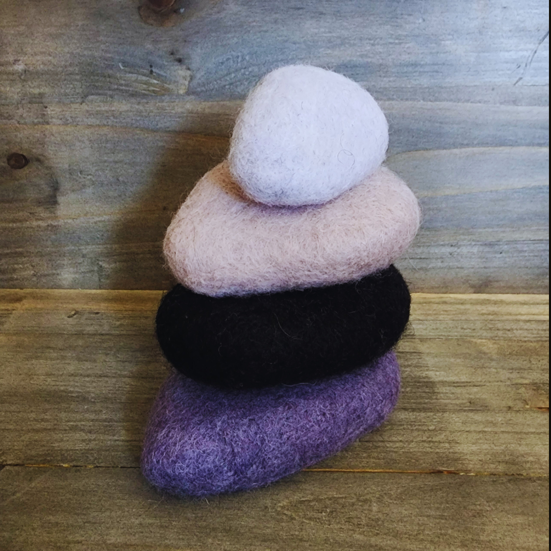 Four handmade wet-felted Zen Stones stacked vertically, from bottom to top: dark purple, black, light pink/beige, and light lavender/white, on a wooden background.