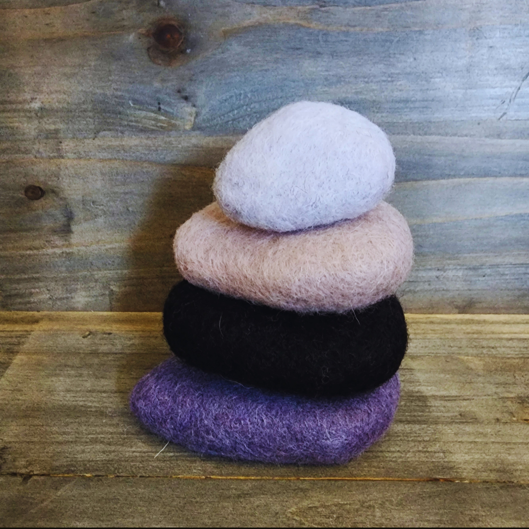 A slightly angled view of four handmade wet-felted Zen Stones stacked, showing textures and colors of dark purple, black, light pink/beige, and light lavender/white, on a rustic wooden background.