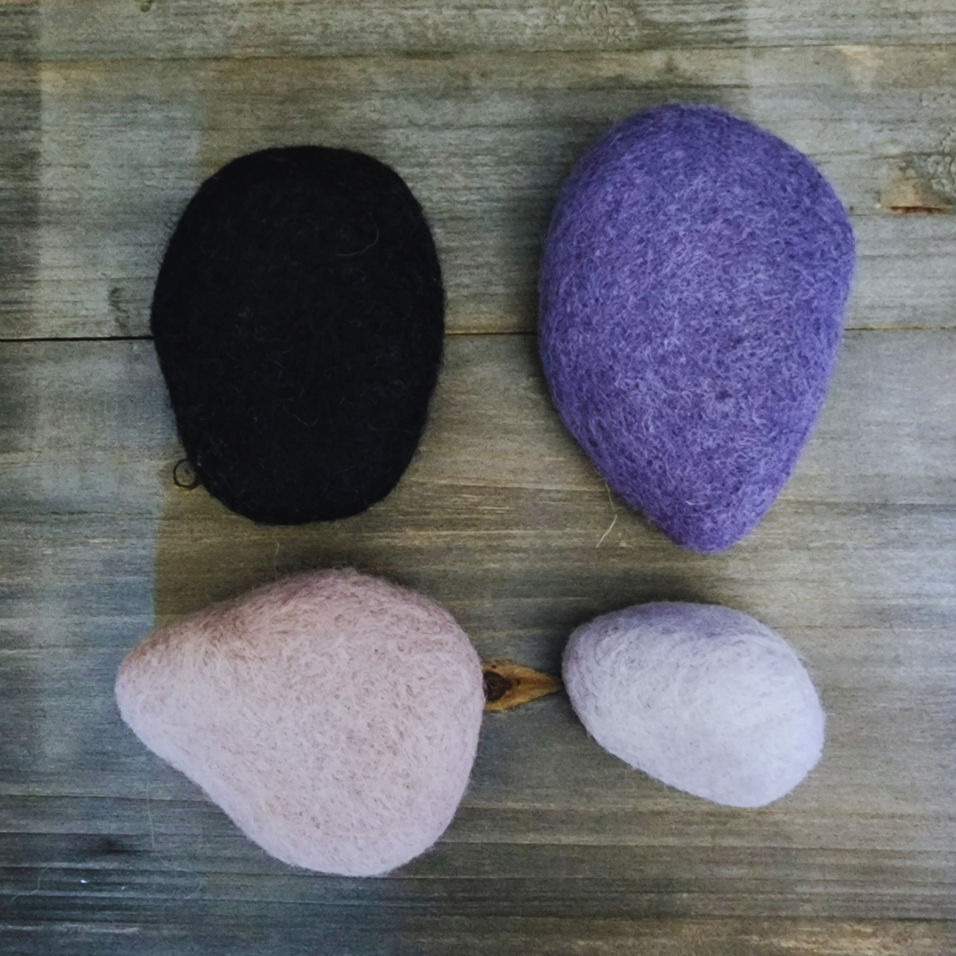 Four handmade wet-felted Zen Stones in an assortment of colors (black, purple, light pink/beige, and light lavender/white), arranged unstacked on a wooden surface.