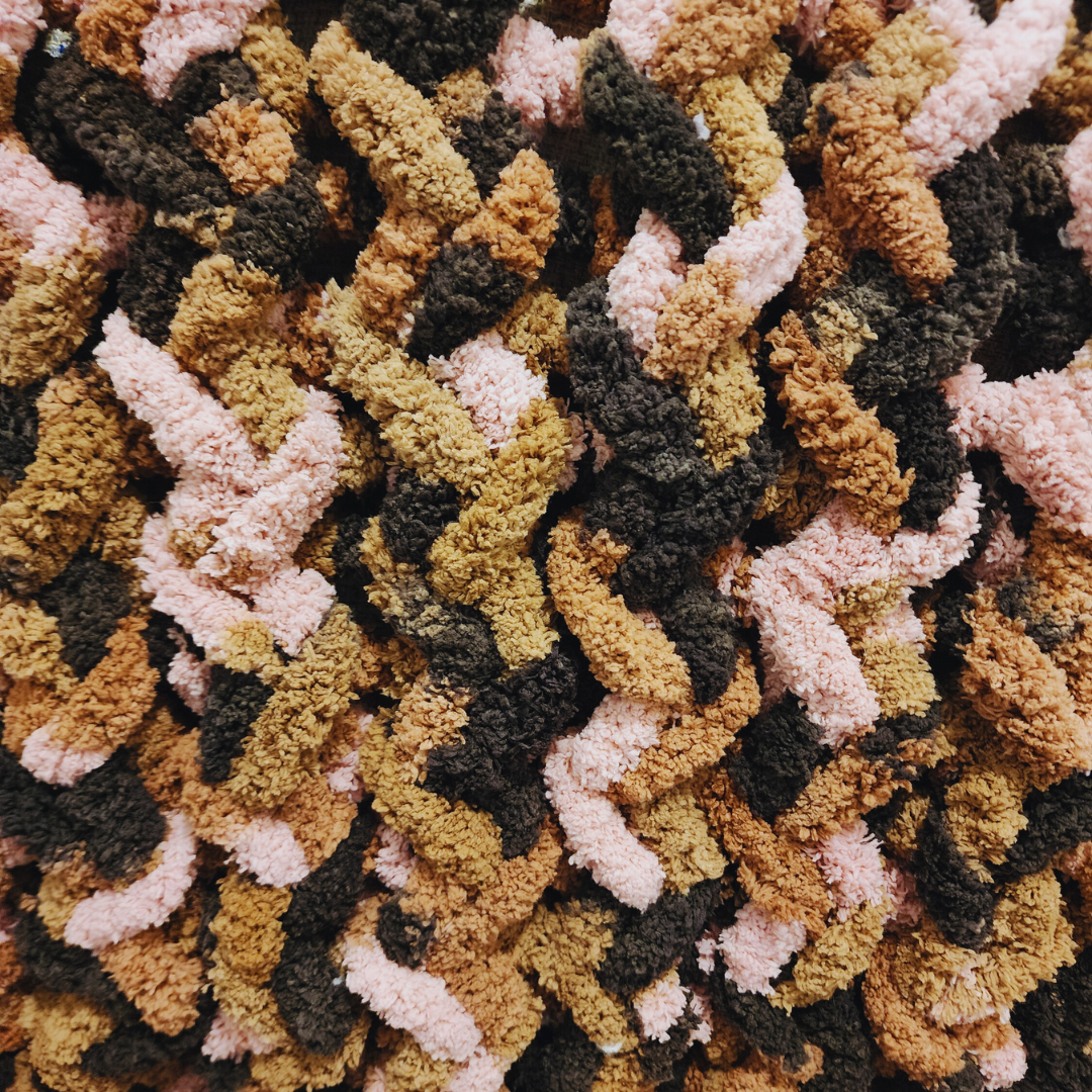 A close-up, overhead view of a handmade chunky knit blanket, showing its soft, textured surface and the zig-zag stitch pattern in brown, pink, and black multicolor yarn.