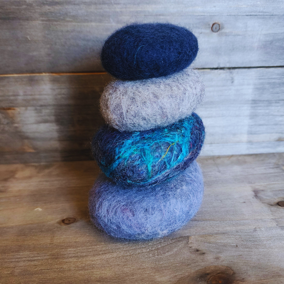 Four handmade wet-felted Zen Stones stacked in a vertical column, from bottom to top: light blue/lavender, dark blue with vivid turquoise and yellow fibers, light gray, and deep navy blue, on a wooden background.
