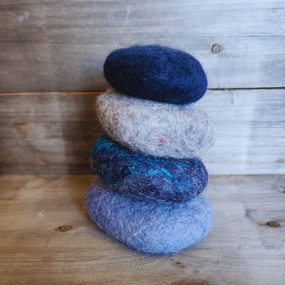 A close-up view of four handmade wet-felted Zen Stones stacked, showing textures and colors of light blue/lavender, dark blue with turquoise threads, light gray, and deep navy blue, against a wooden surface.
