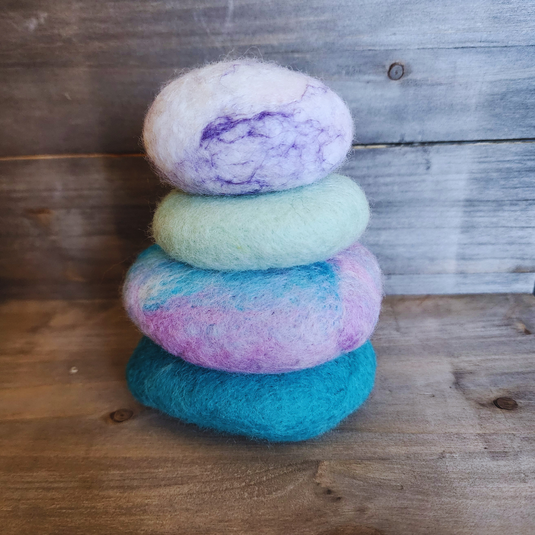 Four handmade wet-felted Zen Stones stacked in a neat column, showing vibrant colors of teal, a pink and blue swirl, mint green, and white with purple accents, against a wooden background.