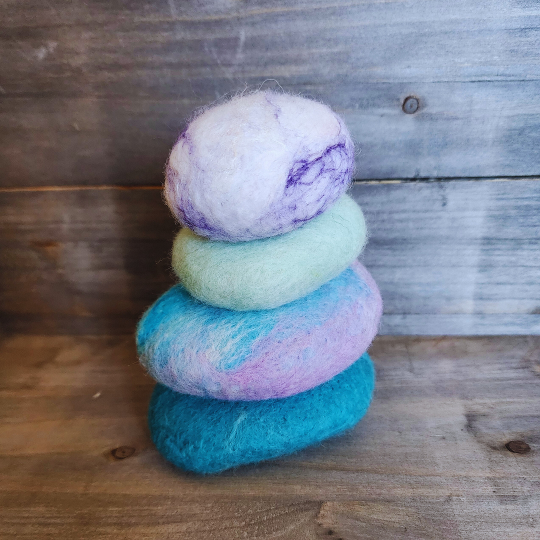 Four handmade wet-felted Zen Stones stacked vertically in pastel colors, from bottom to top: teal, pink and light blue swirl, mint green, and white with purple marbling, on a wooden surface.