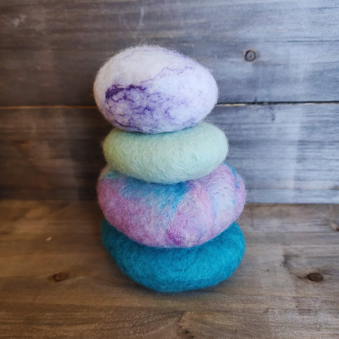 A close-up view of four handmade wet-felted Zen Stones stacked, displaying soft pastel shades of teal, pink and blue swirl, mint green, and white with purple marbling, on a rustic wooden background.