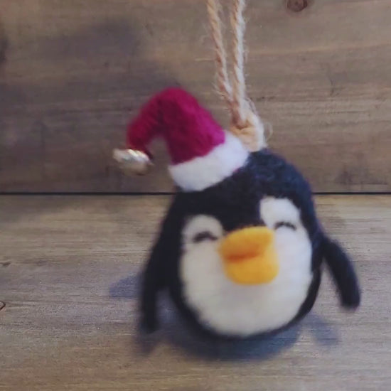 A short video clip showing a hand holding a small, black and white needle-felted penguin ornament that is wearing a red Santa hat with a tiny silver bell. The hand gently turns the ornament to show the front, side, and back, highlighting the fuzzy wool texture, the secure jute string hanger, and the movement of the bell.