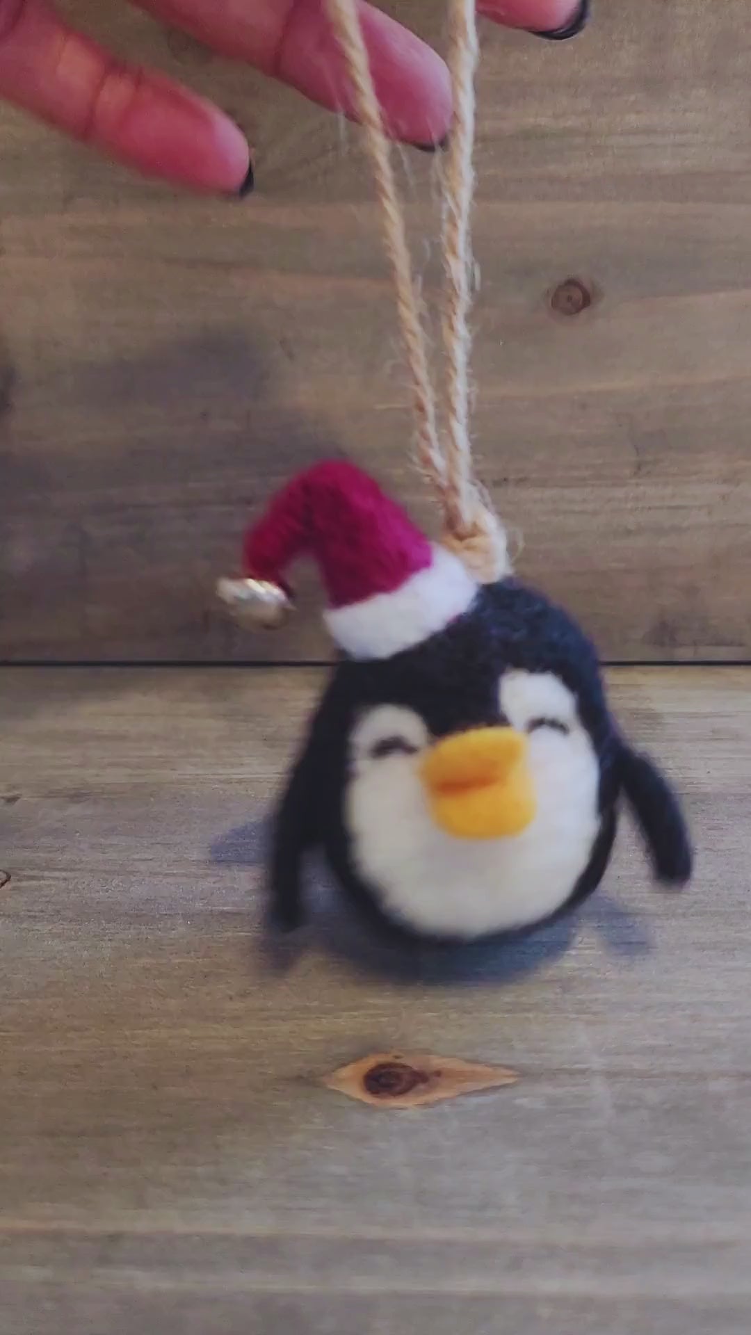 A short video clip showing a hand holding a small, black and white needle-felted penguin ornament that is wearing a red Santa hat with a tiny silver bell. The hand gently turns the ornament to show the front, side, and back, highlighting the fuzzy wool texture, the secure jute string hanger, and the movement of the bell.