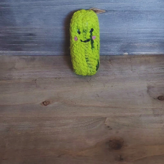 A person’s hand picks up a small, handmade green felted pickle cat toy from a wooden surface and holds it up to the camera. The toy has a sweet, needle-felted smiley face with pink cheeks and dark green "pickle" markings. The person rotates the toy, showing its thick, durable texture from all angles, including the top and bottom, before setting it back down.
