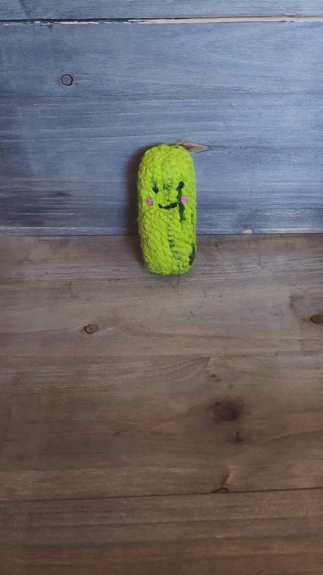 A person’s hand picks up a small, handmade green felted pickle cat toy from a wooden surface and holds it up to the camera. The toy has a sweet, needle-felted smiley face with pink cheeks and dark green "pickle" markings. The person rotates the toy, showing its thick, durable texture from all angles, including the top and bottom, before setting it back down.