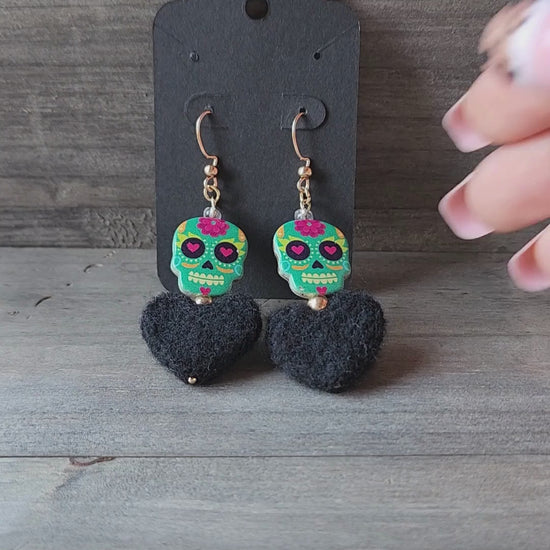 A close-up video of a hand holding a pair of handmade earrings. The video shows the details of the colorful sugar skull beads and the soft, black felt hearts.