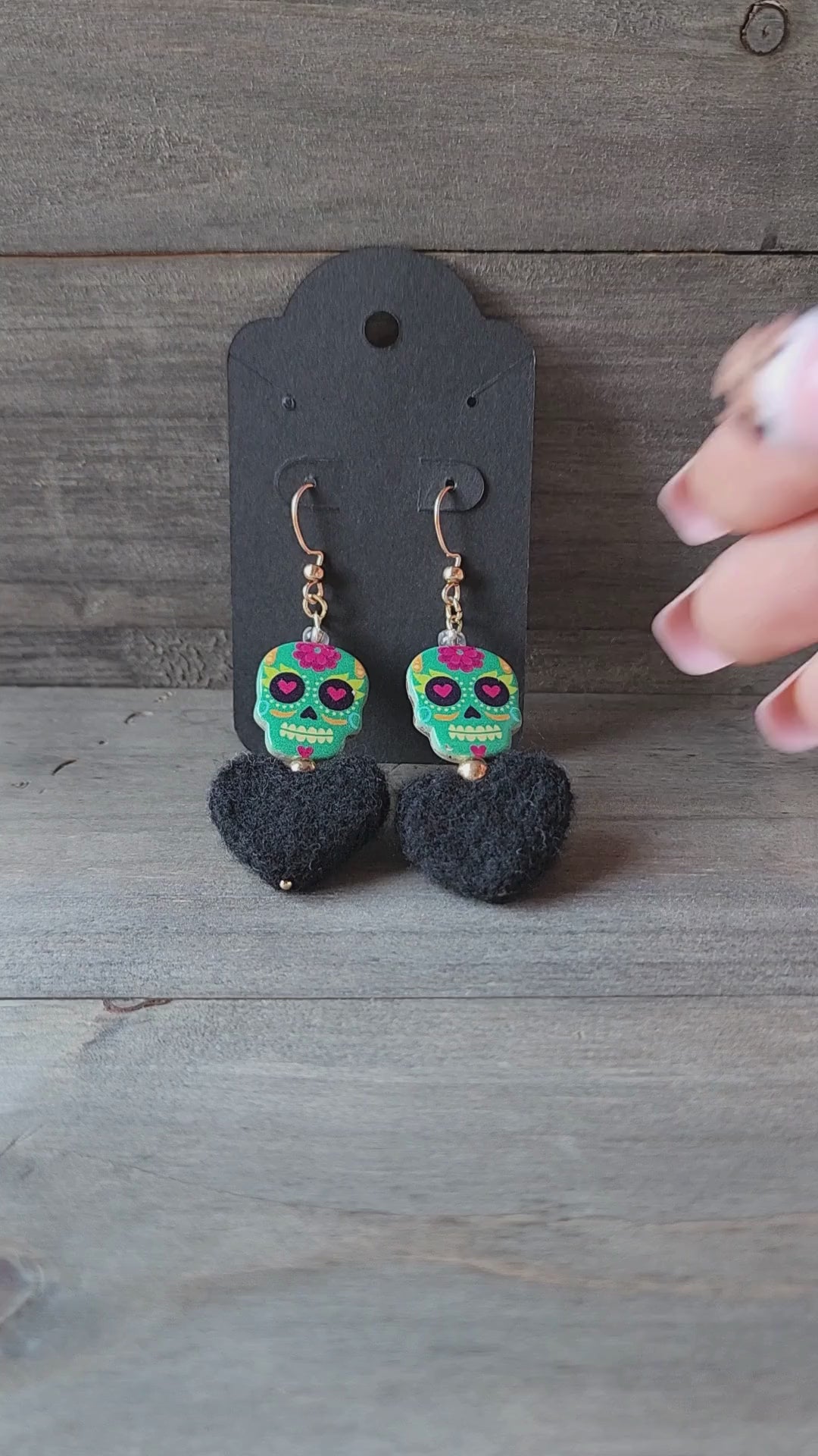 A close-up video of a hand holding a pair of handmade earrings. The video shows the details of the colorful sugar skull beads and the soft, black felt hearts.