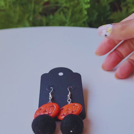 A hand holds up a pair of handmade earrings, featuring an orange jack-o-lantern bead and a black felt ball, to show their size and movement.