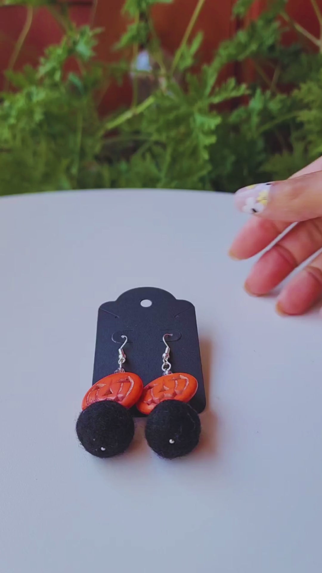 A hand holds up a pair of handmade earrings, featuring an orange jack-o-lantern bead and a black felt ball, to show their size and movement.