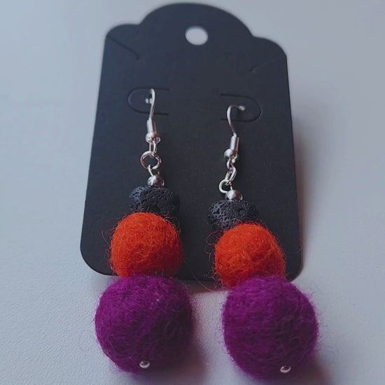 A hand holds up a pair of handmade Halloween earrings, featuring a black lava bead heart with orange and purple felt balls, to show their size and movement.