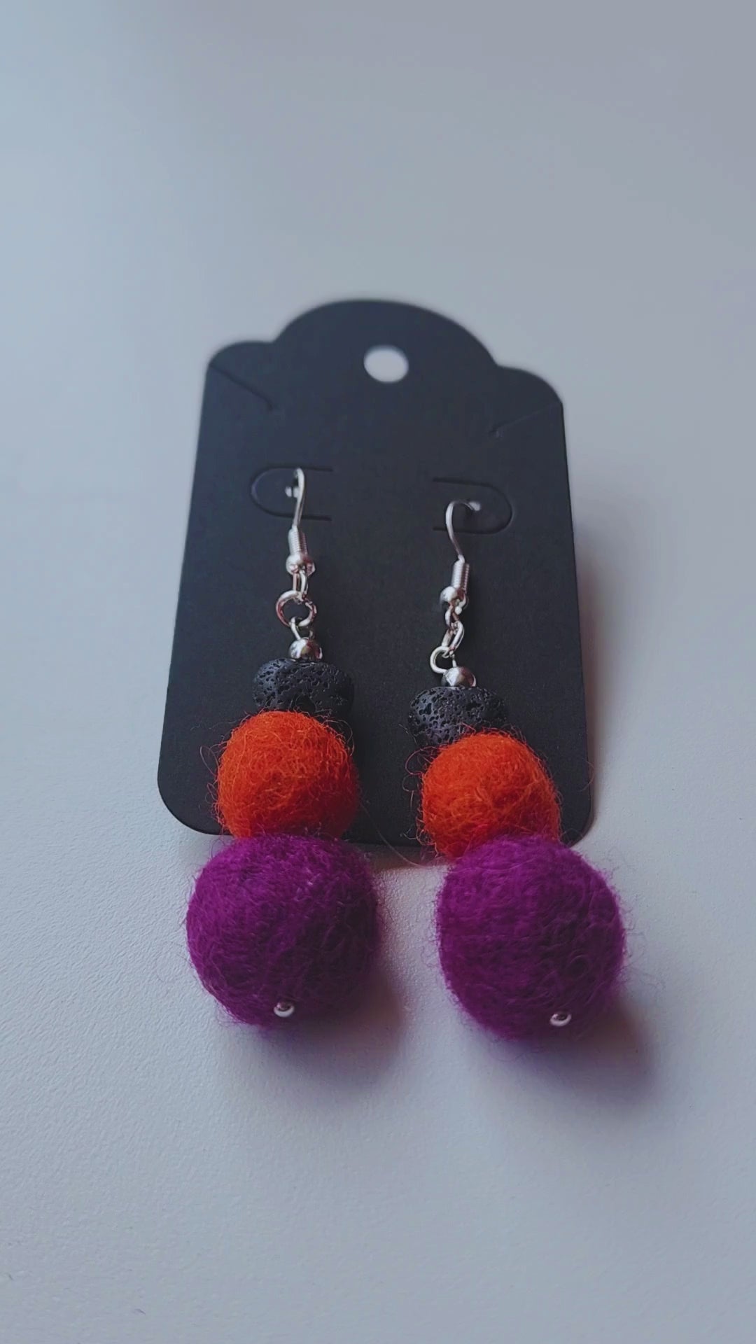 A hand holds up a pair of handmade Halloween earrings, featuring a black lava bead heart with orange and purple felt balls, to show their size and movement.