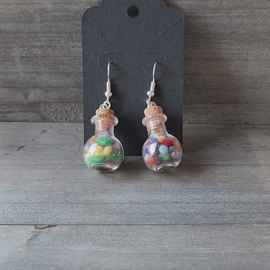 A video of my hand holding up a pair of the handmade potion bottle earrings to show their size and detail. The camera focuses on the tiny glass bottles filled with colorful wool nepps.