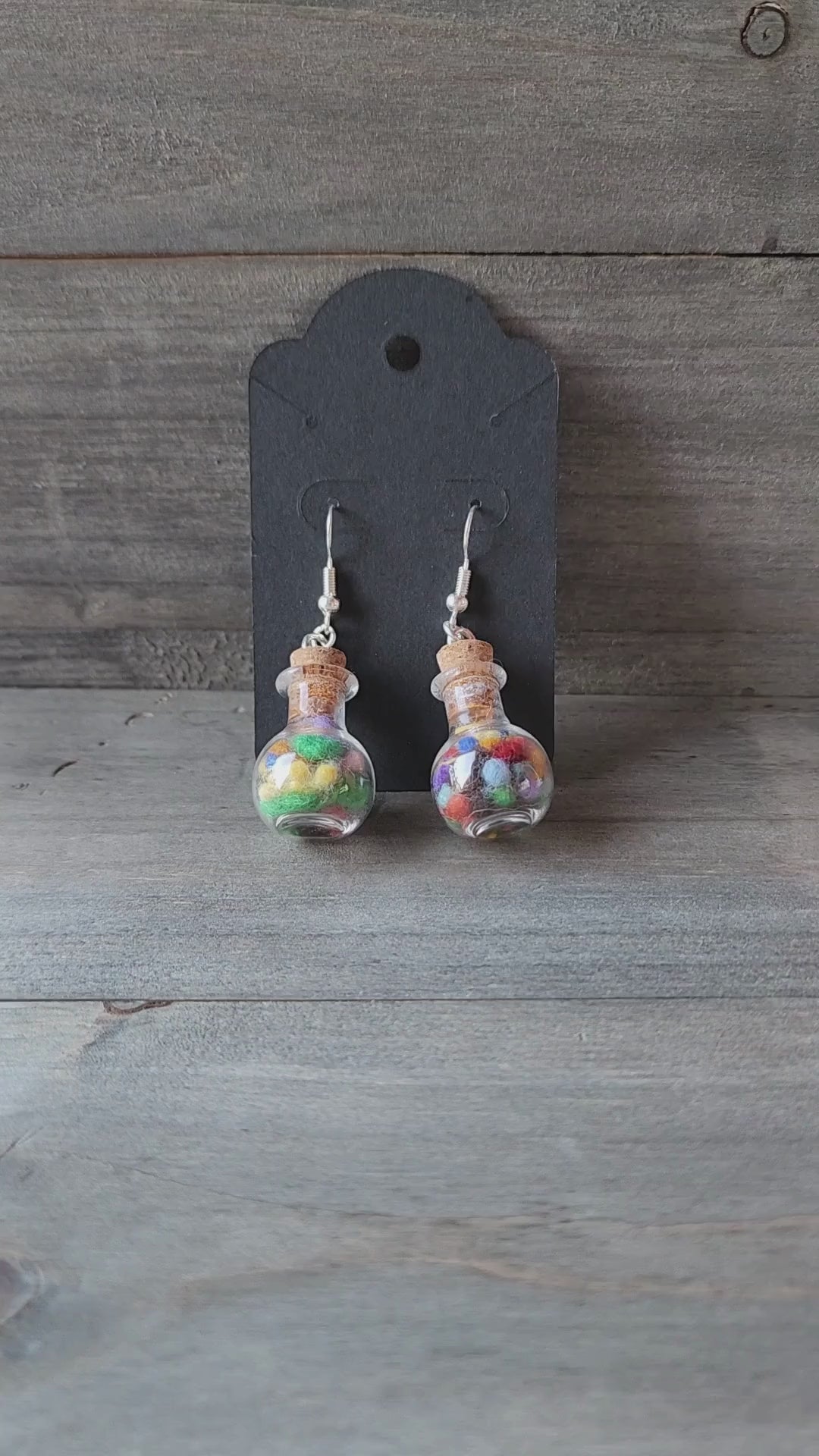 A video of my hand holding up a pair of the handmade potion bottle earrings to show their size and detail. The camera focuses on the tiny glass bottles filled with colorful wool nepps.