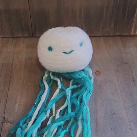 A hand holds up a handmade catnip jellyfish cat toy, showcasing its long, wiggly blue and white yarn tentacles before setting it down on a wooden surface.