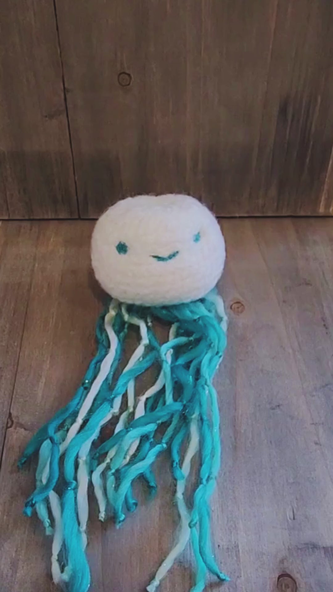 A hand holds up a handmade catnip jellyfish cat toy, showcasing its long, wiggly blue and white yarn tentacles before setting it down on a wooden surface.