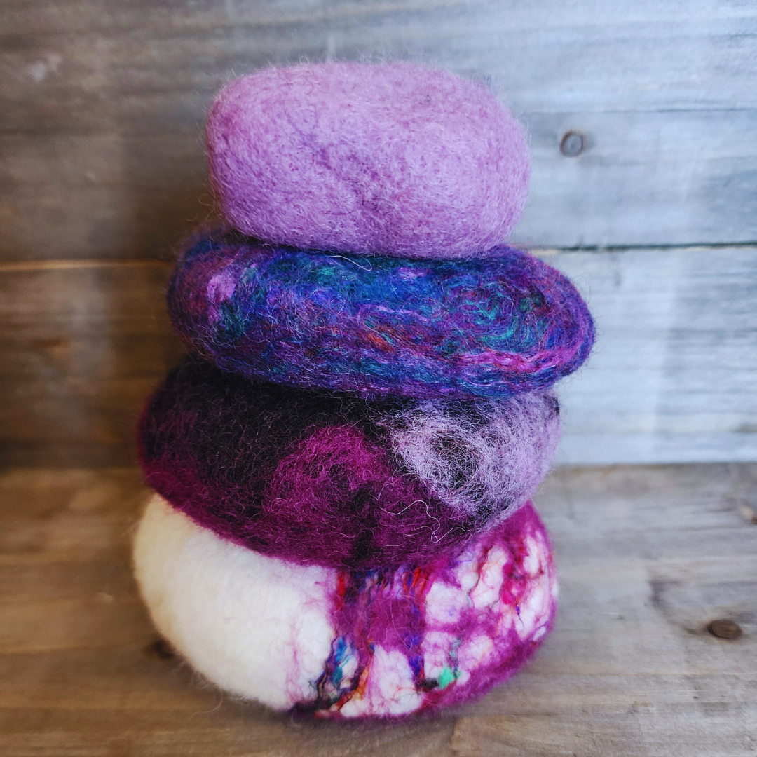 Four handmade wet-felted Zen Stones stacked vertically, featuring vibrant shades of white with purple/magenta marbling, dark purple, mixed blue/purple, and light lavender, on a wooden background.
