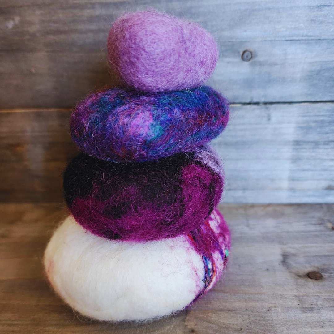 A close-up side view of four handmade wet-felted Zen Stones stacked, showing textures and colors of white with purple/magenta, dark purple, mixed blue/purple, and light lavender, against a wooden surface.